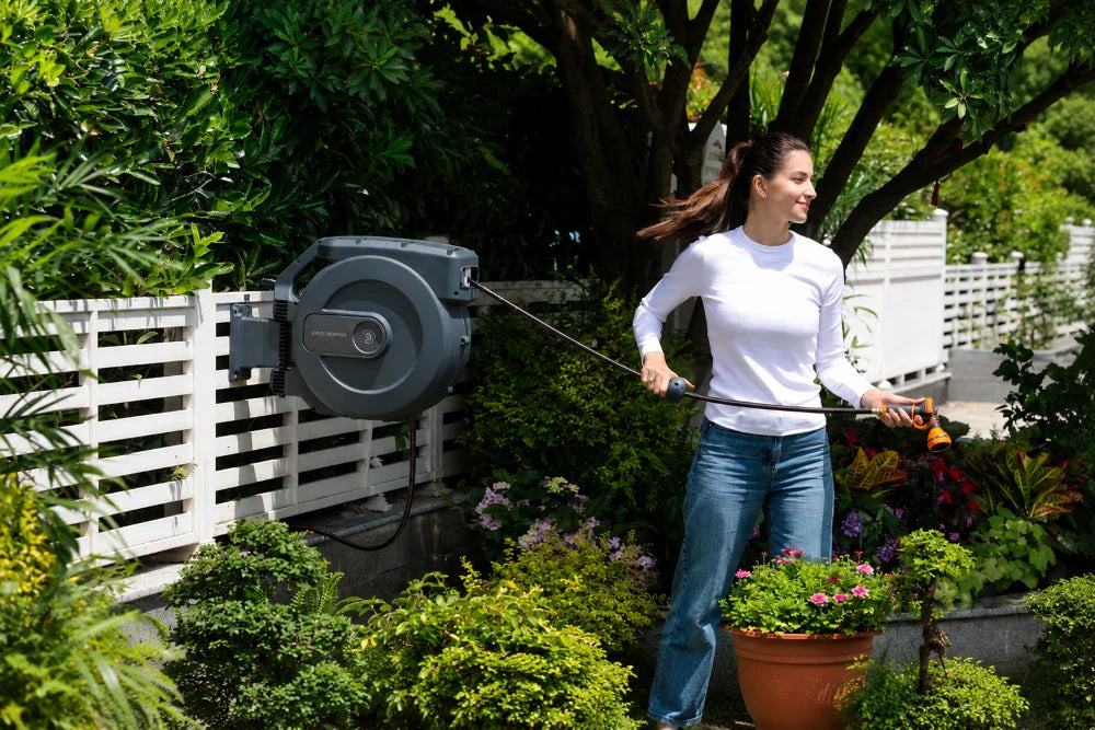 Woman using a wall-mounted retractable garden hose reel in a lush backyard garden.