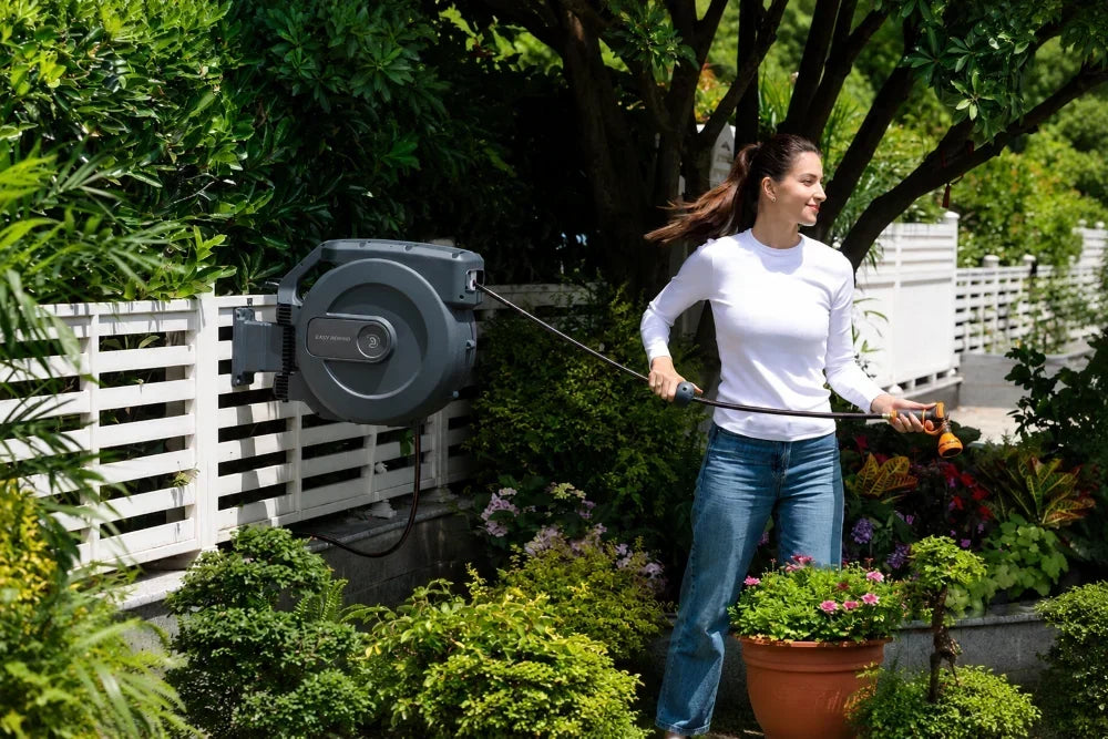 Woman using a wall-mounted retractable garden hose reel in a lush backyard garden.
